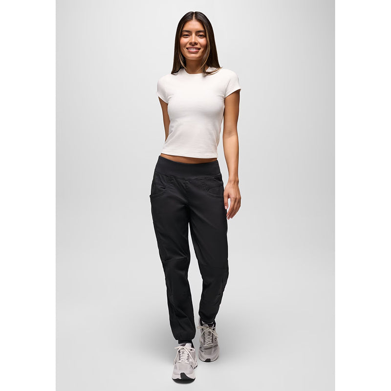 A woman modeling black athletic pants with a white cropped t-shirt and gray sneakers, standing against a light gray studio background.