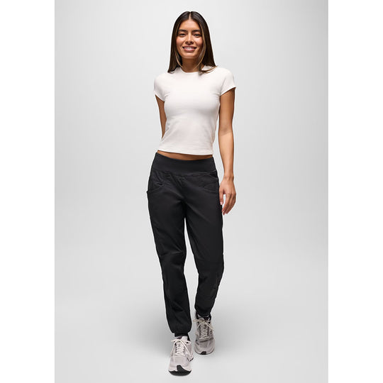 A woman modeling black athletic pants with a white cropped t-shirt and gray sneakers, standing against a light gray studio background.