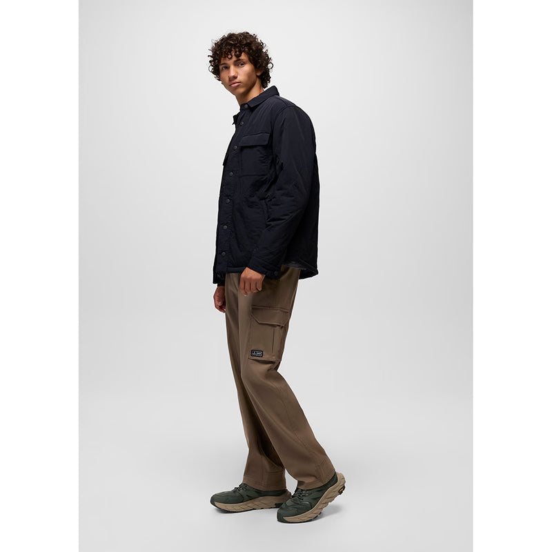 A man wearing a dark navy utility jacket, brown cargo pants, and green hiking shoes, standing in profile against a light gray studio background.