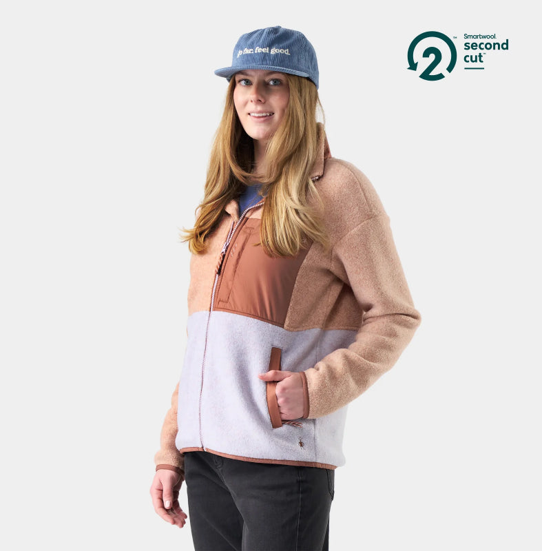 A woman wearing a color-block fleece jacket in beige, brown, and light gray, paired with a blue cap and black pants, set against a plain light gray background. A 'Smartened second cut' logo is visible in the top right corner.