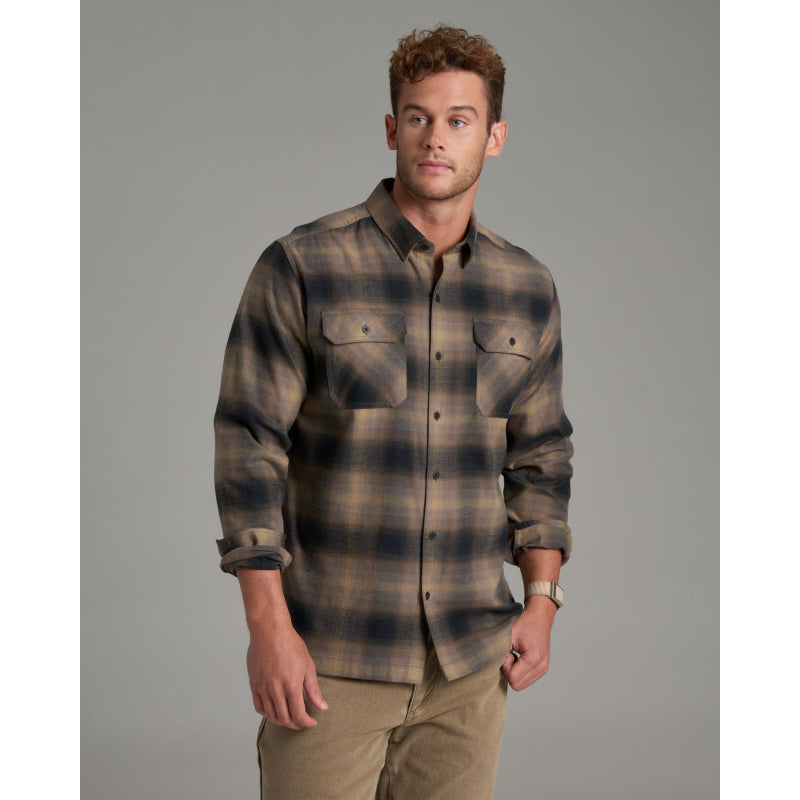 A man wearing a long-sleeve plaid button-up shirt in brown and navy tones, paired with khaki pants, standing against a solid gray studio background.