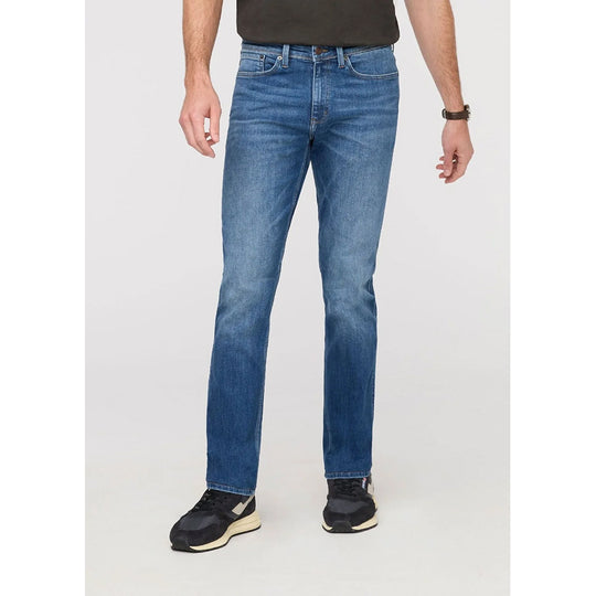 A person wearing blue straight-leg denim jeans, a dark brown t-shirt, and black sneakers, shown from the waist down against a light gray studio background.