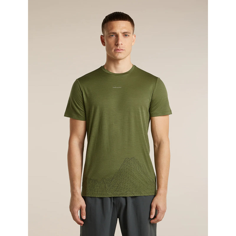A man wearing an olive green athletic t-shirt with a subtle mountain line graphic near the hem, paired with dark gray athletic pants, standing against a solid beige background.
