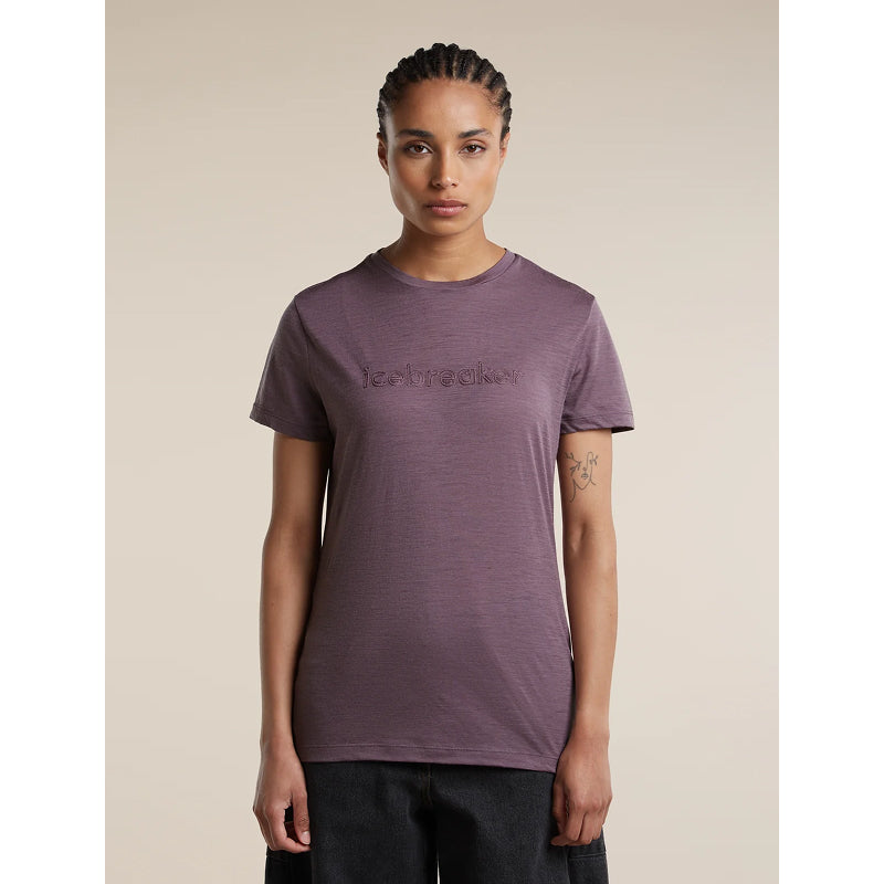 A woman modeling a short-sleeve crewneck t-shirt in a heathered dusty purple color, featuring a tonal embroidered logo on the chest. She is standing against a solid beige background.