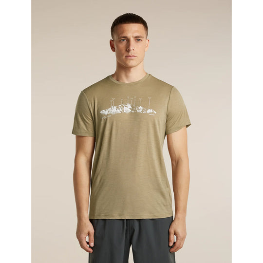 A man wearing a short-sleeve crewneck t-shirt in olive khaki with a white graphic of a mountain and lighthouse on the chest, paired with dark gray athletic pants, set against a solid beige background.