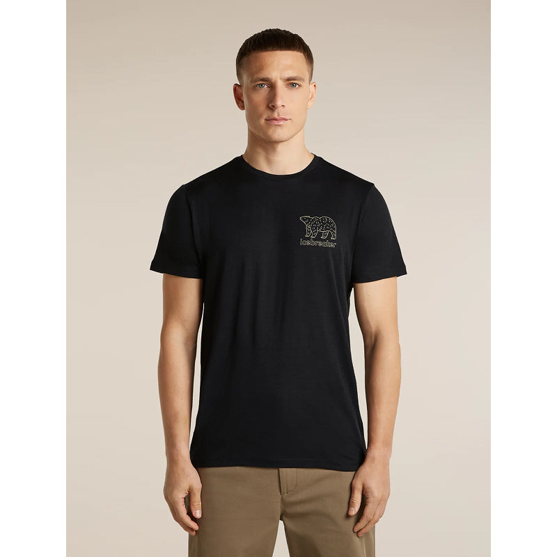A man wearing a black short-sleeve crewneck t-shirt with a small dark embroidered polar bear graphic and the word 'indivisibility' on the left chest, paired with khaki pants, against a solid beige background.