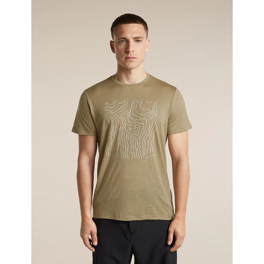 A man wearing a short-sleeve crewneck t-shirt in olive khaki with a white topographic line graphic on the chest, paired with black pants, set against a solid beige studio background.