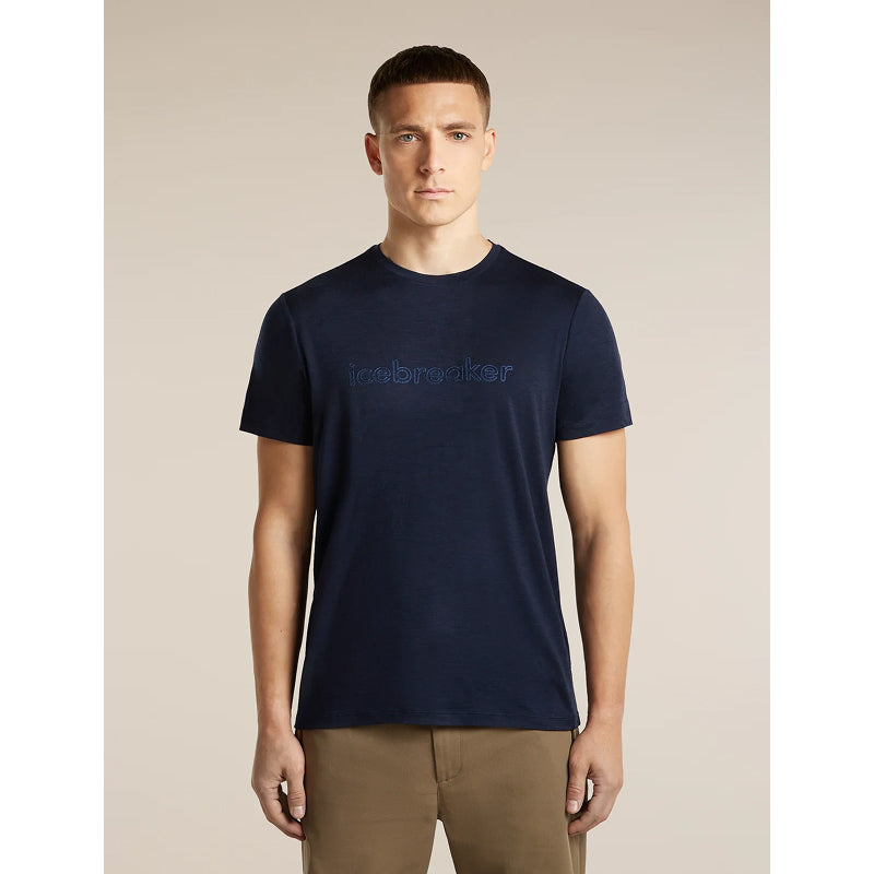 A man wearing a navy blue crewneck t-shirt with 'icebreaker' embroidered in tonal blue lettering across the chest, paired with khaki pants, set against a solid beige background.