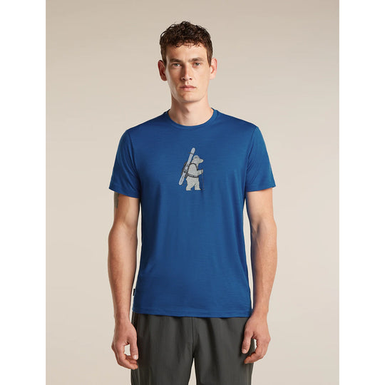A man wearing a royal blue crewneck t-shirt with a small white graphic of a bear carrying a ski pole on the chest, paired with dark gray shorts, set against a plain beige studio background.