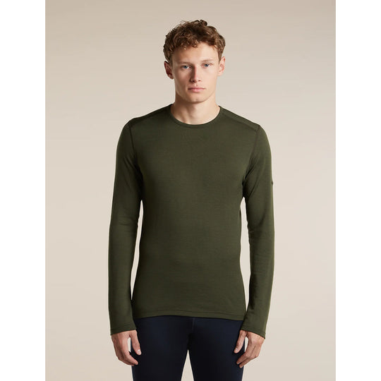 A man wearing a dark olive green long-sleeve crewneck shirt, paired with dark navy pants, standing against a solid beige studio background.