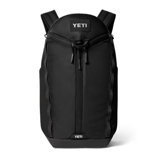 A black YETI backpack shown from the front on a clean white background. The backpack features a top zip closure, a central vertical zipper, side compression straps, and the YETI brand name visible on the top and bottom patches.